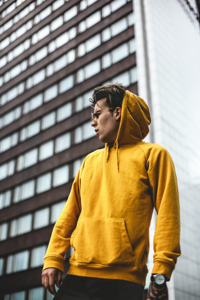 Stylish man poses in yellow hoodie against urban building backdrop.